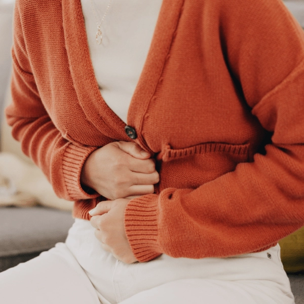 Woman in an orange sweater sitting and clutching her stomach in discomfort, depicting digestive side effects such as nausea or abdominal pain.