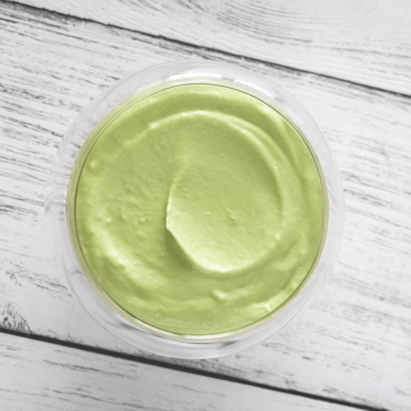 A glass bowl filled with smooth, light green avocado mayo, viewed from above on a white wooden surface.