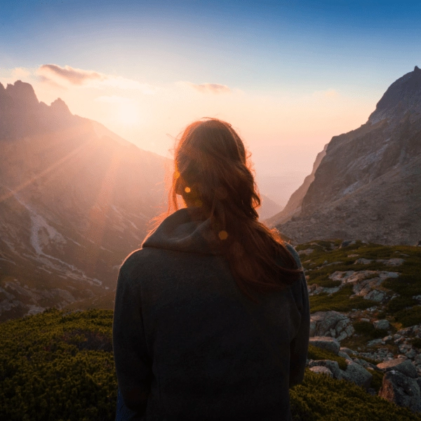A woman stands on a mountain ridge at sunrise, looking out over a glowing valley with sun rays breaking through the peaks. The scene symbolizes reflection, new beginnings, and the empowering journey toward healing and wholeness.