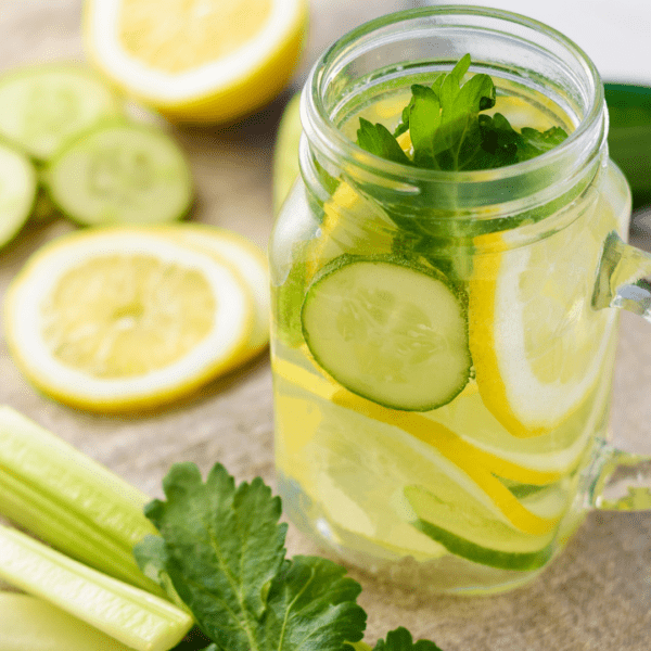 A mason jar filled with cucumber, lemon slices, and herbs in water, with fresh cucumber and lemon slices on a table around it.