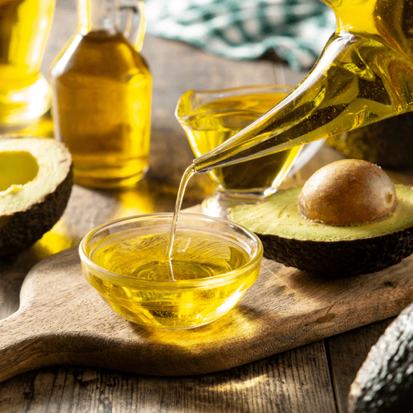 Golden avocado oil being poured from a glass container into a small clear bowl, with fresh avocados in the background.