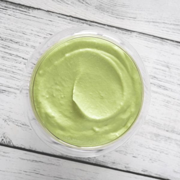 A glass bowl filled with smooth, light green avocado mayo, viewed from above on a white wooden surface.
