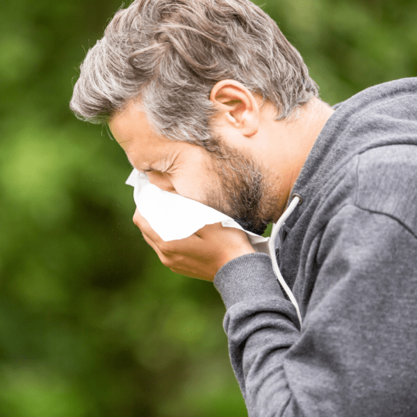 A middle-aged man sneezing into a tissue outdoors, wearing a gray hoodie, surrounded by greenery, indicating allergy symptoms.