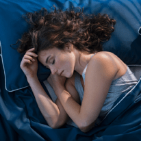 A young woman with curly hair resting comfortably on her side in a dark blue bed, looking relaxed and deeply asleep.