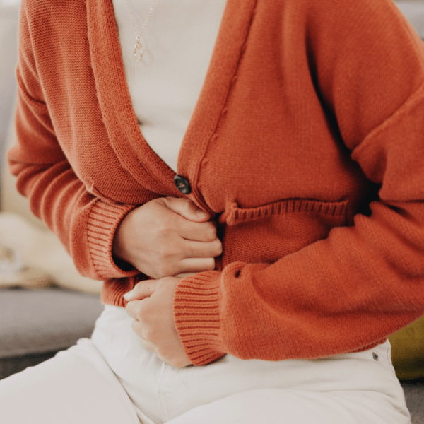 Woman in an orange sweater sitting and clutching her stomach in discomfort, depicting digestive side effects such as nausea or abdominal pain.