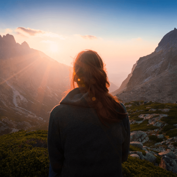 A woman stands on a mountain ridge at sunrise, looking out over a glowing valley with sun rays breaking through the peaks. The scene symbolizes reflection, new beginnings, and the empowering journey toward healing and wholeness.