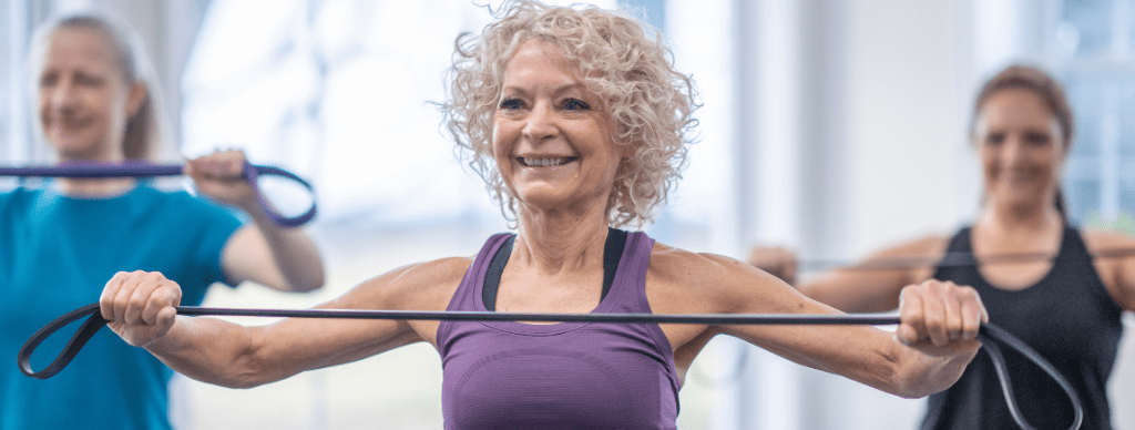 Older woman smiling and exercising in a group fitness class, lifting a resistance band, demonstrating the benefits of staying active and strong during midlife.