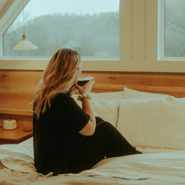 Woman sitting in bed with tousled blonde hair, holding a warm drink and gazing out a window on a cozy morning.