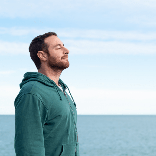 Man in a green hoodie standing by the ocean with eyes closed, taking a deep, peaceful breath.
