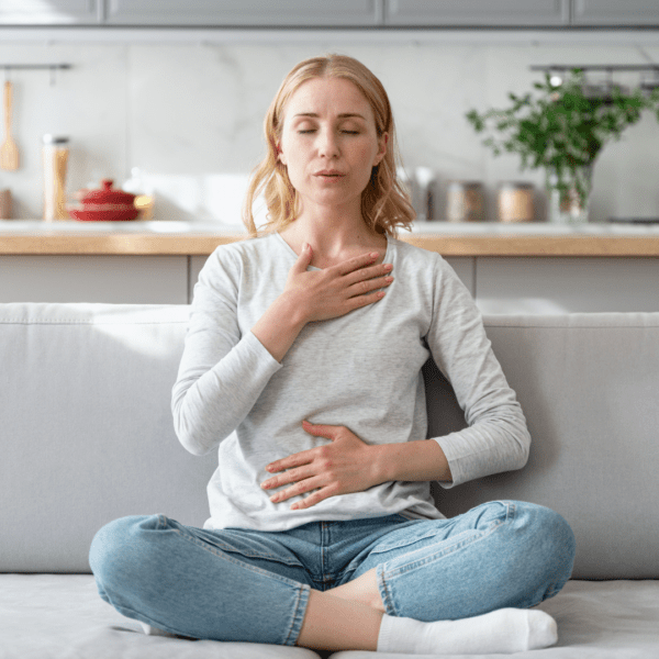 A woman sits cross-legged on a light gray couch with eyes closed, practicing deep breathing. One hand is placed on her chest and the other on her abdomen, demonstrating diaphragmatic breathing.