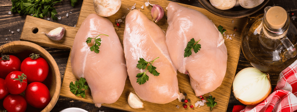 Three raw chicken breasts garnished with fresh parsley are arranged on a wooden cutting board. Surrounding the board are fresh ingredients including tomatoes, garlic, onion, mushrooms, a bottle of olive oil, and leafy herbs.