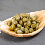 A wooden spoon filled with fresh, green capers, placed on a dark slate background with coarse salt sprinkled nearby.