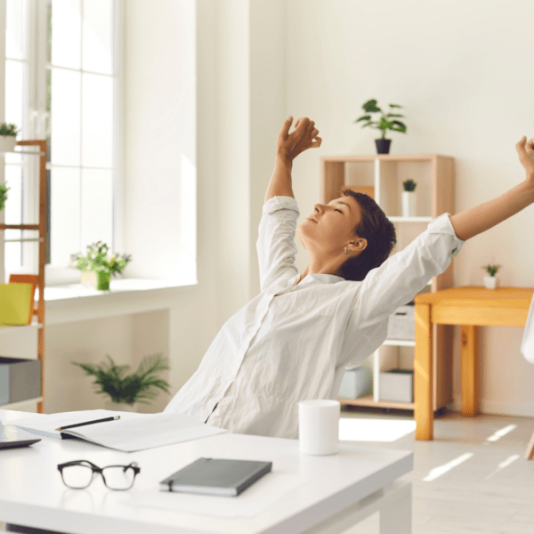 A woman sits at a white desk in a bright, modern home office, stretching with arms raised and eyes closed in a peaceful expression, suggesting a moment of breathwork or relaxation during the workday. 