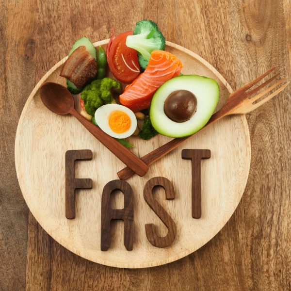 A wooden plate with healthy foods like salmon, avocado, egg, and vegetables arranged to resemble a clock, with the word "FAST" written in wooden letters below.