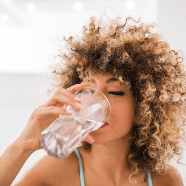 A woman with curly hair drinking a glass of water in a brightly lit room.