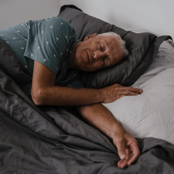 An older man sleeping comfortably on his side in bed, covered in a soft gray blanket, with a relaxed expression on his face.