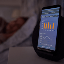 A smartphone on a bedside table displaying a sleep tracking app with graphs and statistics, while a person sleeps in the background.