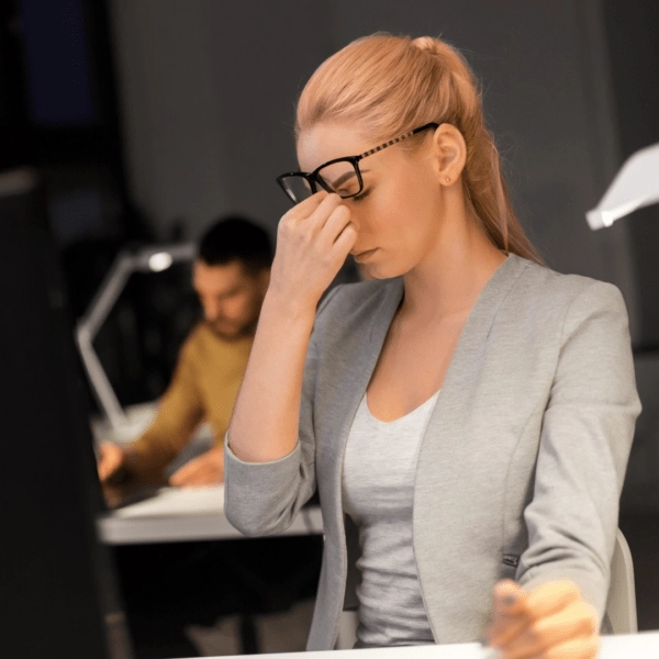 A professional woman wearing glasses, sitting in an office, rubbing her forehead with a tired expression, indicating exhaustion and sleep deprivation.
