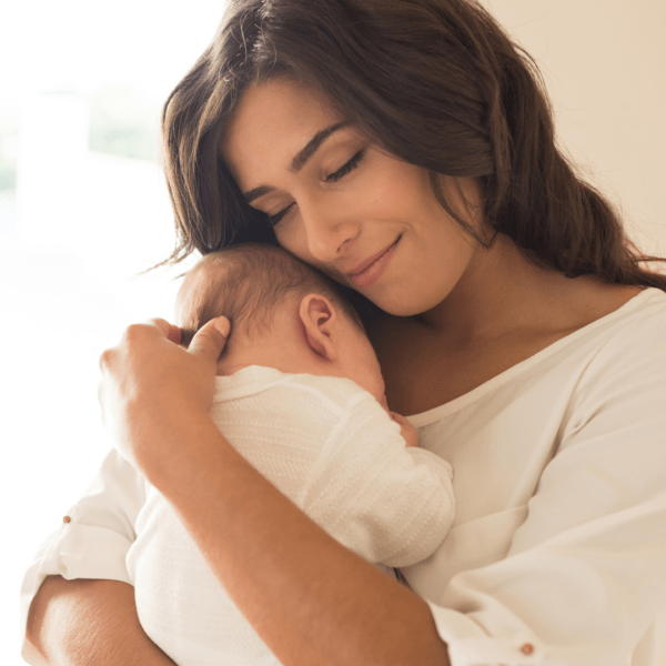 A young woman lovingly cradles a sleeping newborn baby on her chest, conveying warmth and maternal affection in a softly lit environment.