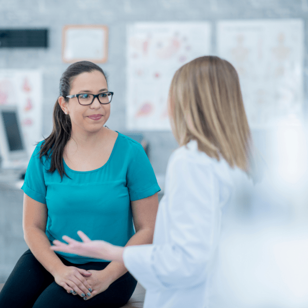 A female healthcare provider in a white coat converses with a female patient wearing glasses and a teal shirt in a clinical setting. They are seated and engaged in a friendly, focused discussion.