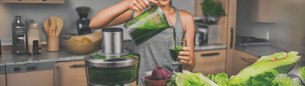 A person in a grey tank top pours a glass of fresh green juice from a blender pitcher, surrounded by fresh vegetables like beets, lettuce, and carrots on a kitchen counter.