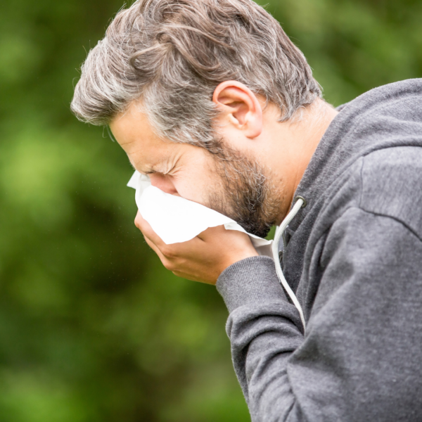 A middle-aged man sneezing into a tissue outdoors, wearing a gray hoodie, surrounded by greenery, indicating allergy symptoms.