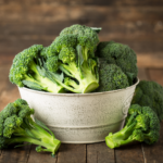 A rustic white metal bowl filled with fresh green broccoli, placed on a wooden table with a few loose broccoli florets.