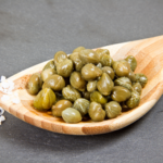 A wooden spoon filled with fresh, green capers, placed on a dark slate background with coarse salt sprinkled nearby.