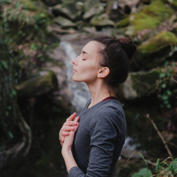 A woman with eyes closed standing in a forest, hands over her heart, practicing mindful breathing.