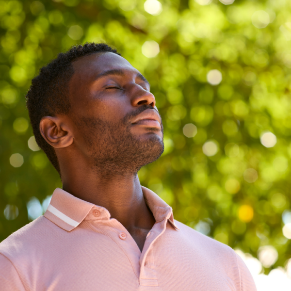 A man in a pink polo shirt standing outdoors with eyes closed, taking a deep breath under bright green trees.