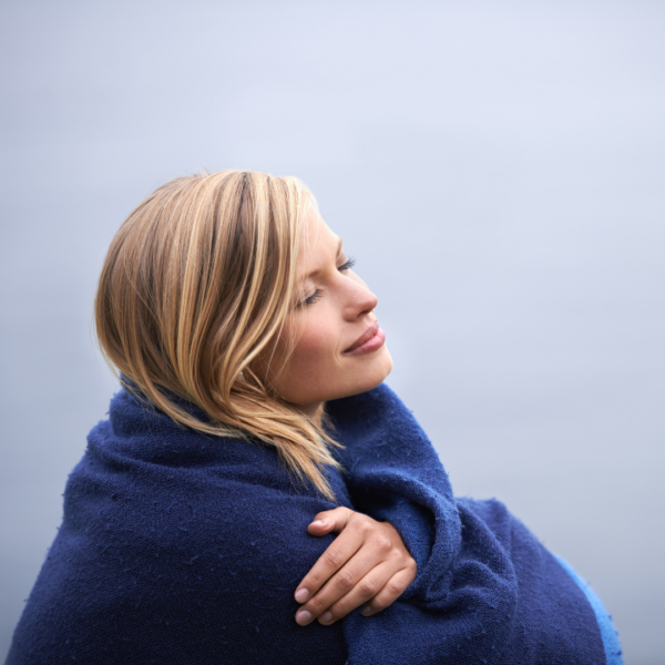 Peaceful woman wrapped in a blue blanket, eyes closed and smiling softly while facing a calm gray sky.