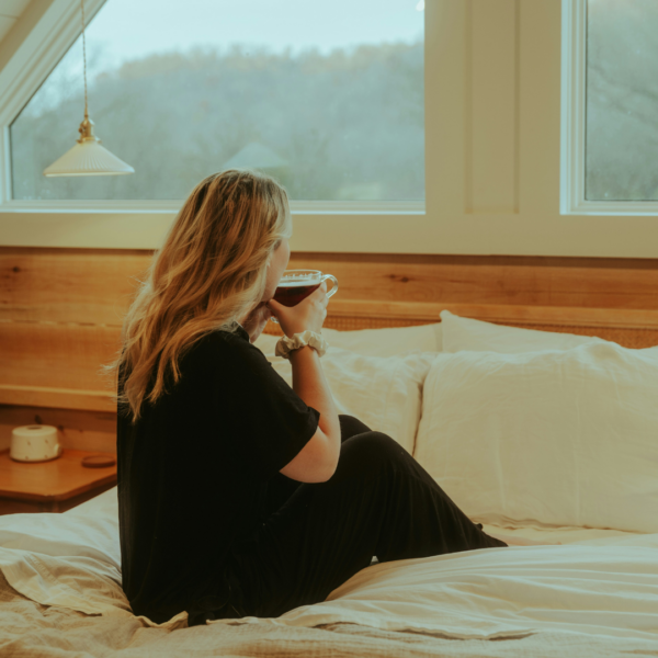 Woman sitting in bed with tousled blonde hair, holding a warm drink and gazing out a window on a cozy morning.