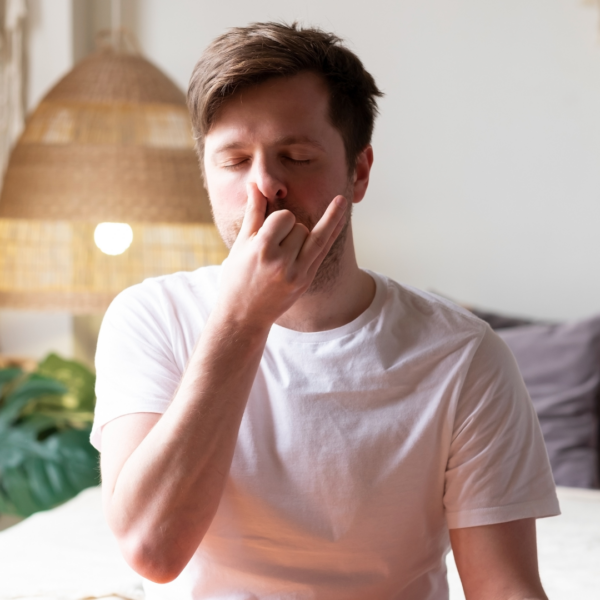 Man in a white t-shirt practicing alternate nostril breathing (nadi shodhana) with his eyes closed at home.