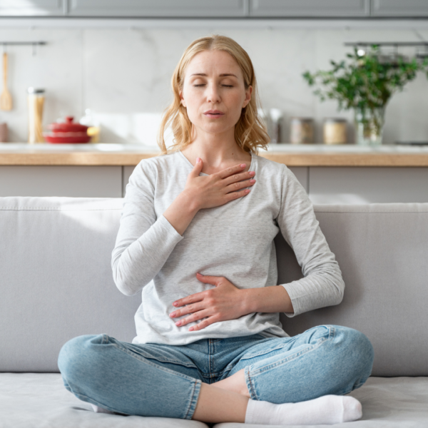A woman sits cross-legged on a light gray couch with eyes closed, practicing deep breathing. One hand is placed on her chest and the other on her abdomen, demonstrating diaphragmatic breathing.