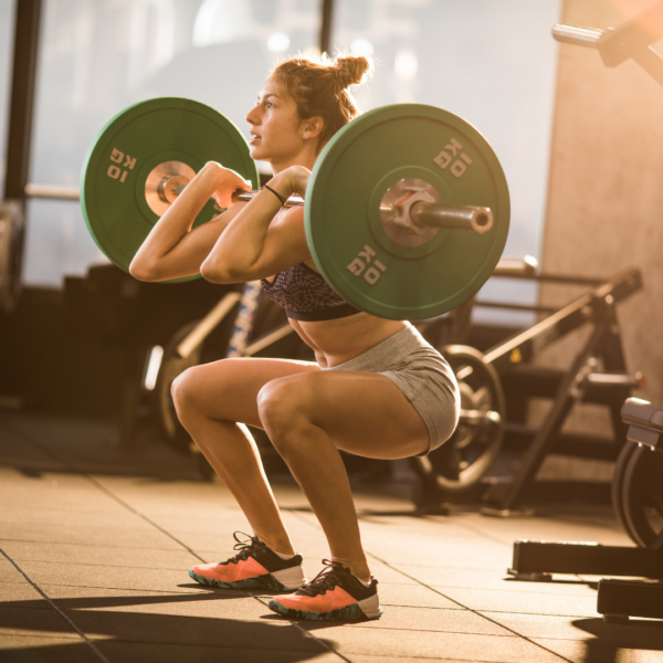 A woman performs a front squat with a loaded barbell in a gym, focusing her breath and posture. She is in a low squat position with elbows forward, lifting weights marked 10 KG, surrounded by gym equipment in a sunlit training space.