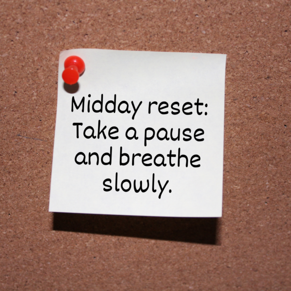 A white sticky note pinned to a corkboard with a red pushpin. The note reads: 'Midday reset: Take a pause and breathe slowly.' The handwriting is casual and clear, offering a gentle reminder for mindful breathing.