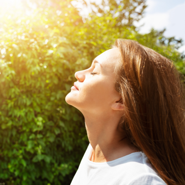 Peaceful woman standing outdoors with closed eyes, basking in sunlight and nature, illustrating calmness, breathwork, or emotional healing.