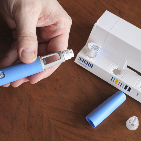 Hand holding a blue GLP-1 injection pen, preparing it for use, with medication packaging and needle caps on a wooden table.
