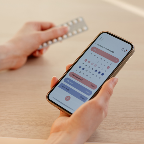 A close-up of a smartphone displaying a menstrual cycle tracking app with a calendar view, held by a person who is also holding a blister pack of pills.