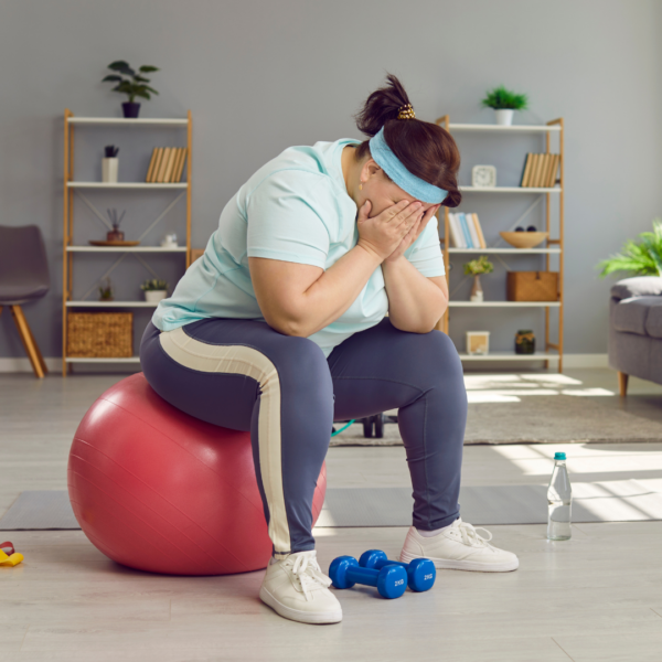 Overweight woman in workout clothes sitting on an exercise ball with her face in her hands, appearing frustrated or discouraged by lack of weight loss progress.