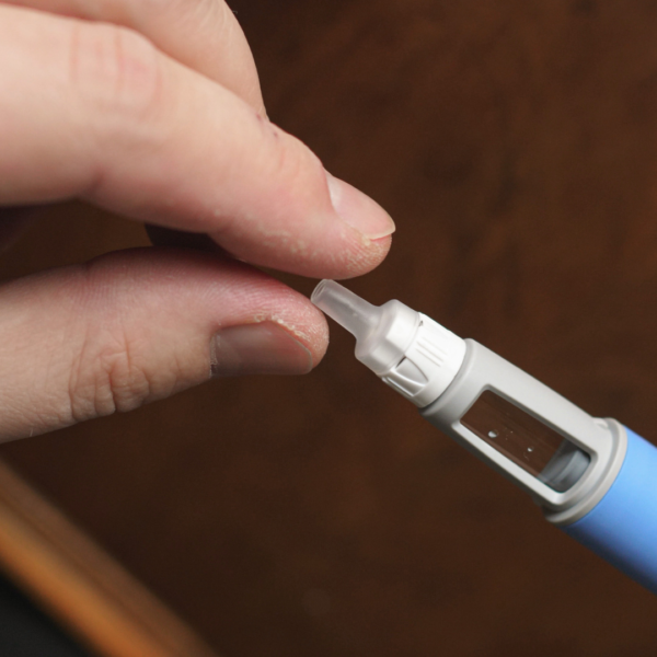 Close-up of fingers attaching a needle cap to a blue GLP-1 injection pen, demonstrating safe medication preparation.