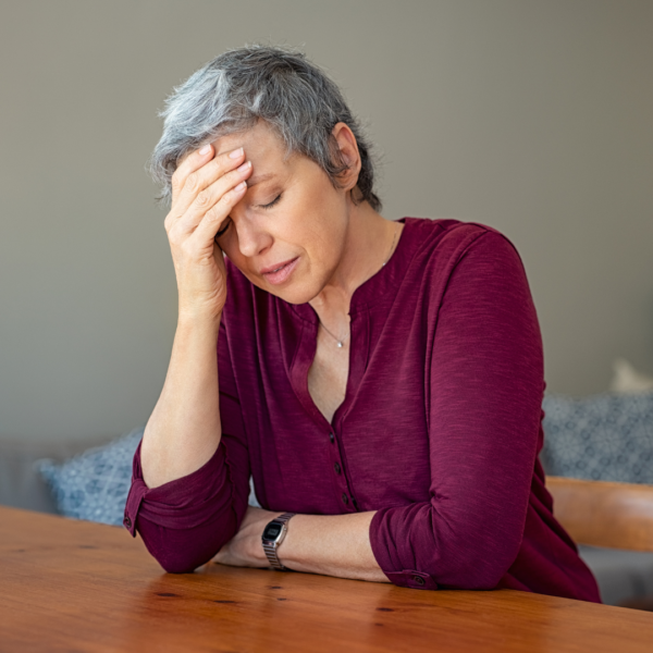 Mature woman with short gray hair sitting at a table with her hand to her forehead, showing fatigue or stress, common during hormonal transitions.