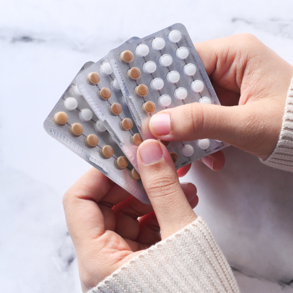 Hands holding several blister packs of oral contraceptive pills, each with white and brown tablets, on a light surface.