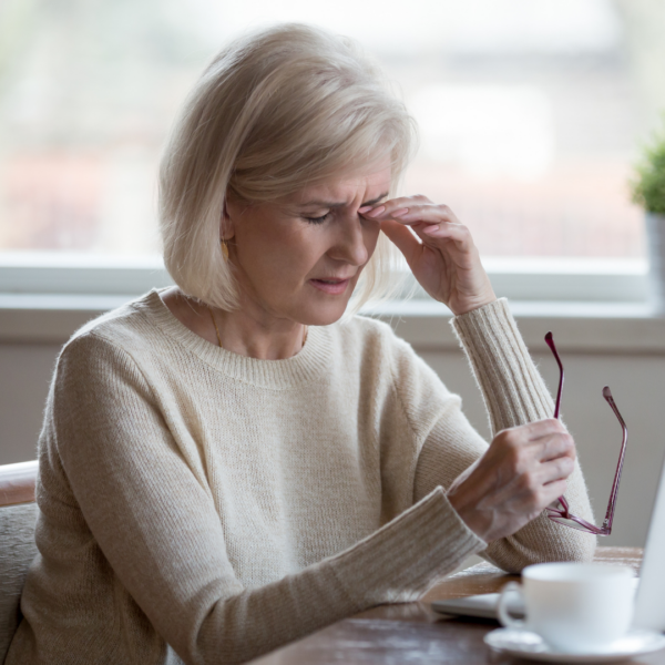 Blonde woman in a cozy sweater sitting at a table with a laptop and coffee, rubbing her forehead as if experiencing a headache or brain fog.