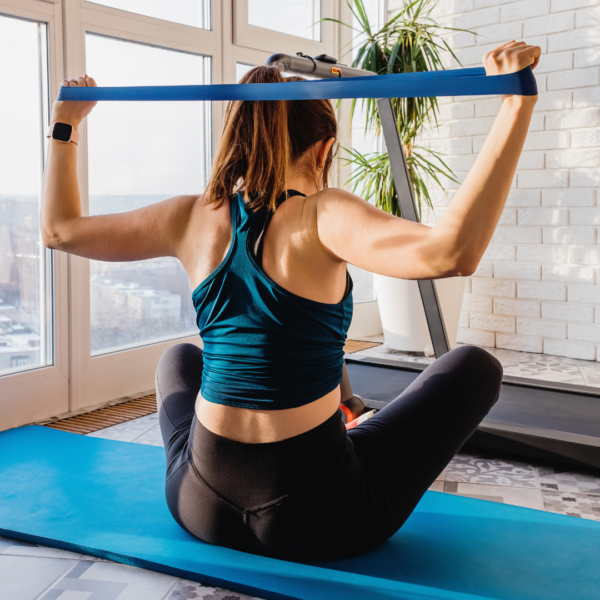 Woman sitting on a yoga mat at home, performing resistance exercises with a blue resistance band, promoting muscle preservation during weight loss.
