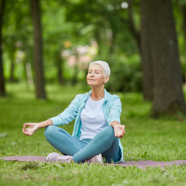 Older woman with short gray hair sitting cross-legged on a yoga mat in a park, meditating with eyes closed and a peaceful expression, illustrating mindfulness and self-care in nature.