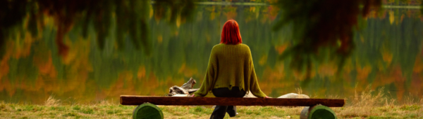 A person with bright red hair sits alone on a wooden bench facing a peaceful lake, surrounded by trees with autumn-colored leaves.