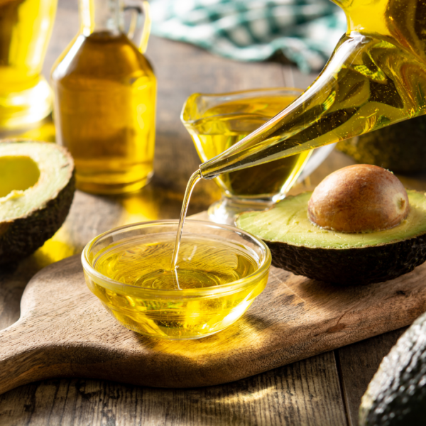 Golden avocado oil being poured from a glass container into a small clear bowl, with fresh avocados in the background.