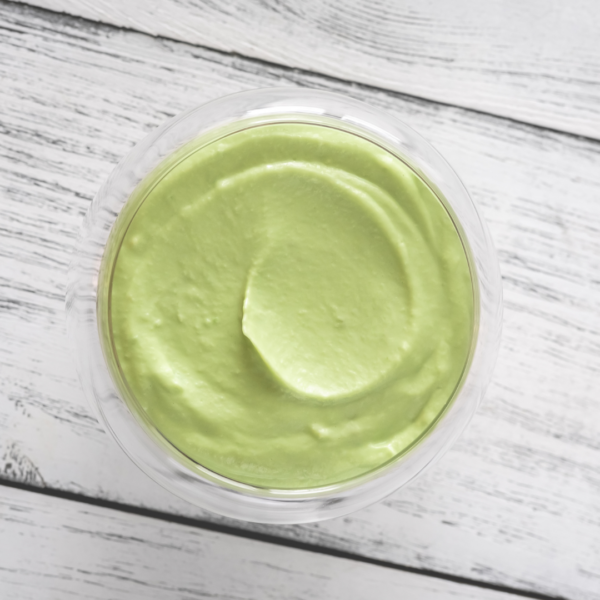 A glass bowl filled with smooth, light green avocado mayo, viewed from above on a white wooden surface.