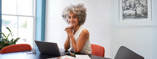 Smiling older woman with curly gray hair sitting at a desk with a laptop in a bright office.